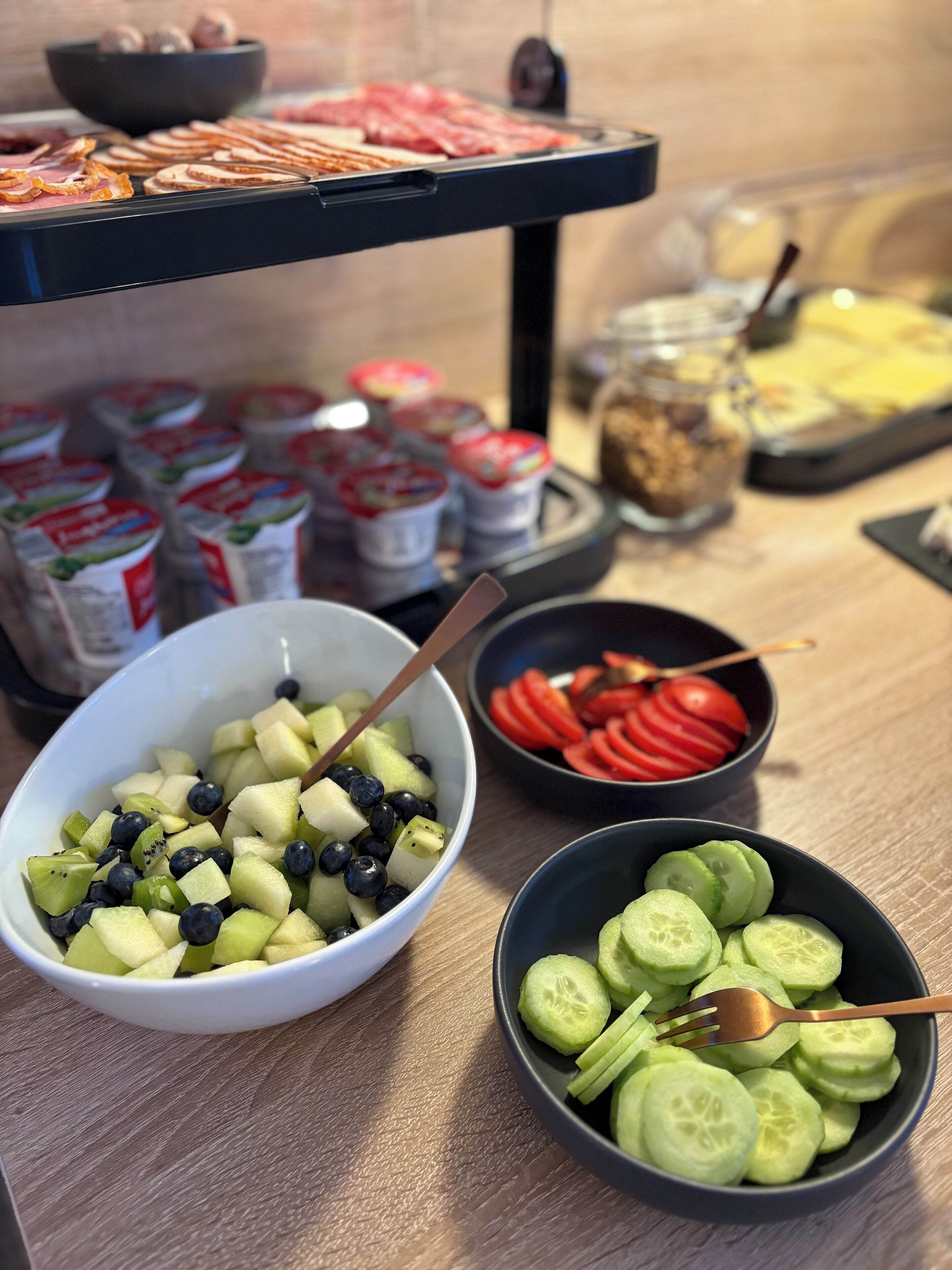 A buffet table with various food items including sliced cucumbers, tomatoes, and a fruit bowl with apples and blueberries.