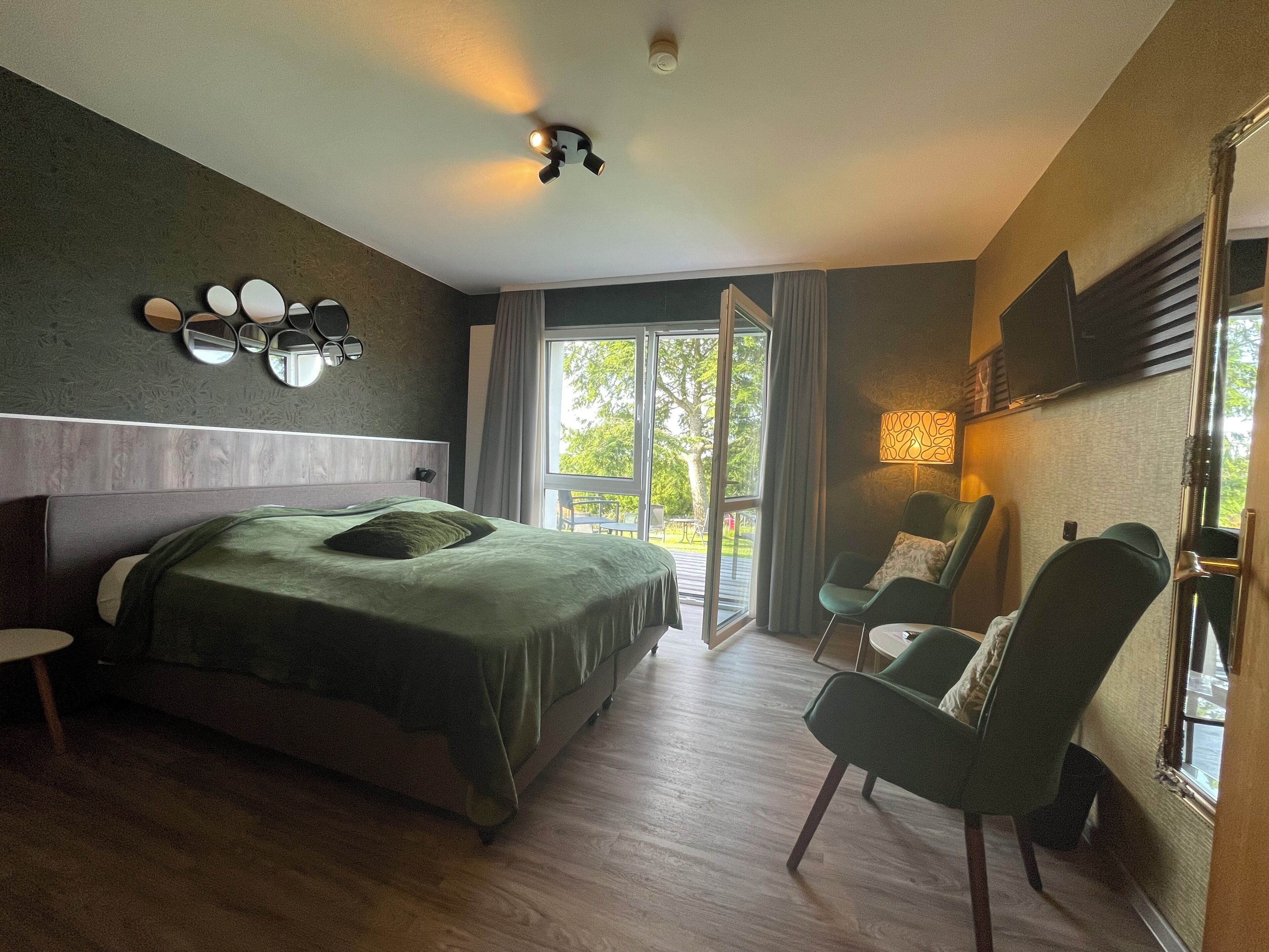 A modern hotel room with a large bed, a seating area with chairs, a TV mounted on the wall, and a balcony with a view of trees.