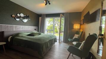 A modern hotel room with a large bed, a seating area with chairs, a TV mounted on the wall, and a balcony with a view of trees.