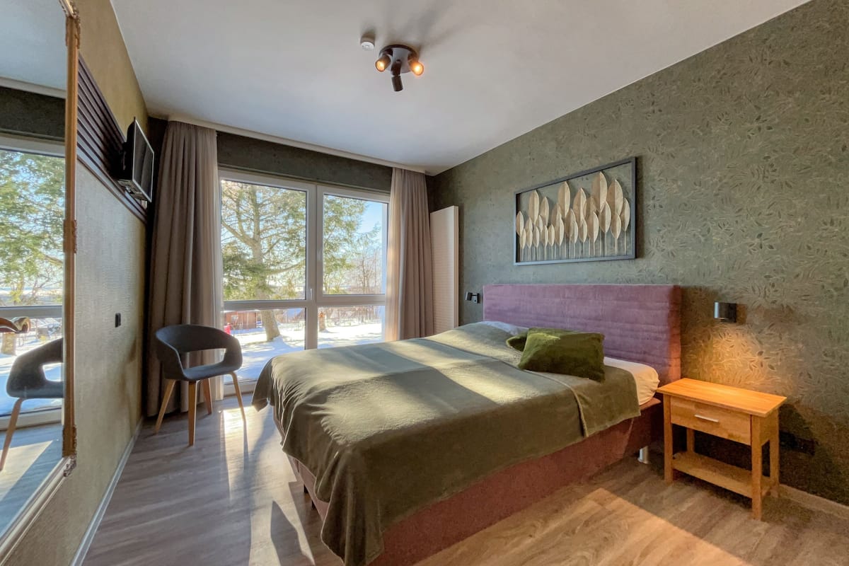 Une chambre à coucher moderne avec un grand lit, une table de chevet, une chaise et une fenêtre donnant sur des arbres.