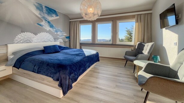 A modern bedroom with a large bed, a sitting area with a chair, a TV mounted on the wall, and large windows offering a view of the outdoors. - Berghotel Hohegeiß (Braunlage)