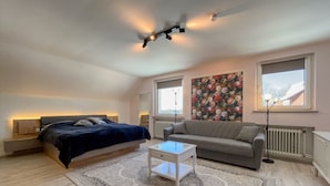 A modern bedroom with a bed, a grey sofa, a small white table, a patterned rug, and a floral wall art. - Berghotel Hohegeiß (Braunlage)