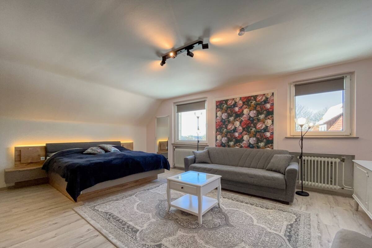 Une chambre moderne avec un lit, un canapé gris, une petite table blanche, un tapis à motifs et une décoration murale florale.