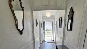 Een gang met een groot, versierd spiegel aan de linkerkant, een donkere kast aan de rechterkant en een deur aan het einde die naar buiten leidt.