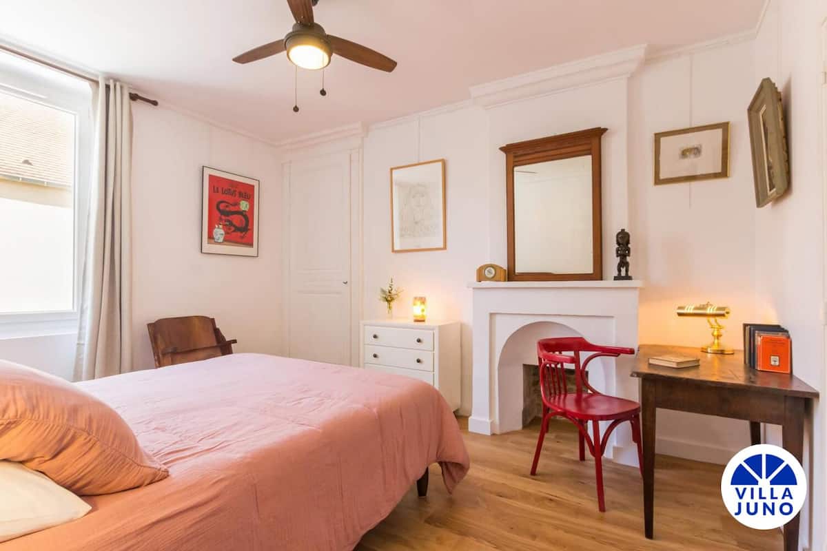 A bedroom with a bed, a desk, a red chair, a fireplace, and a ceiling fan.