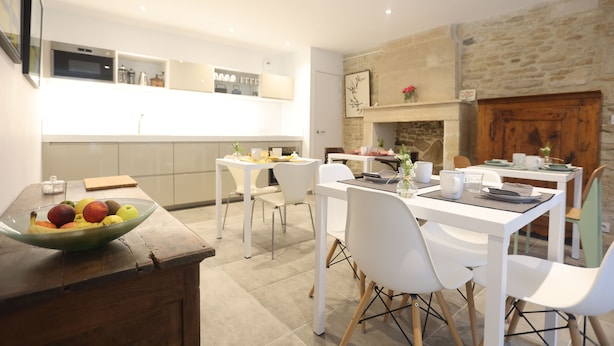 A modern kitchen with white cabinetry, a stone fireplace, and a dining area with white chairs and a table set for two. - Villa Juno (Saint-Aubin-sur-Mer)