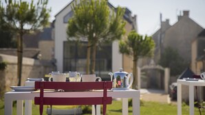 A table set outdoors with a teapot and cups, surrounded by greenery and a house in the background. - Villa Juno (Saint-Aubin-sur-Mer)