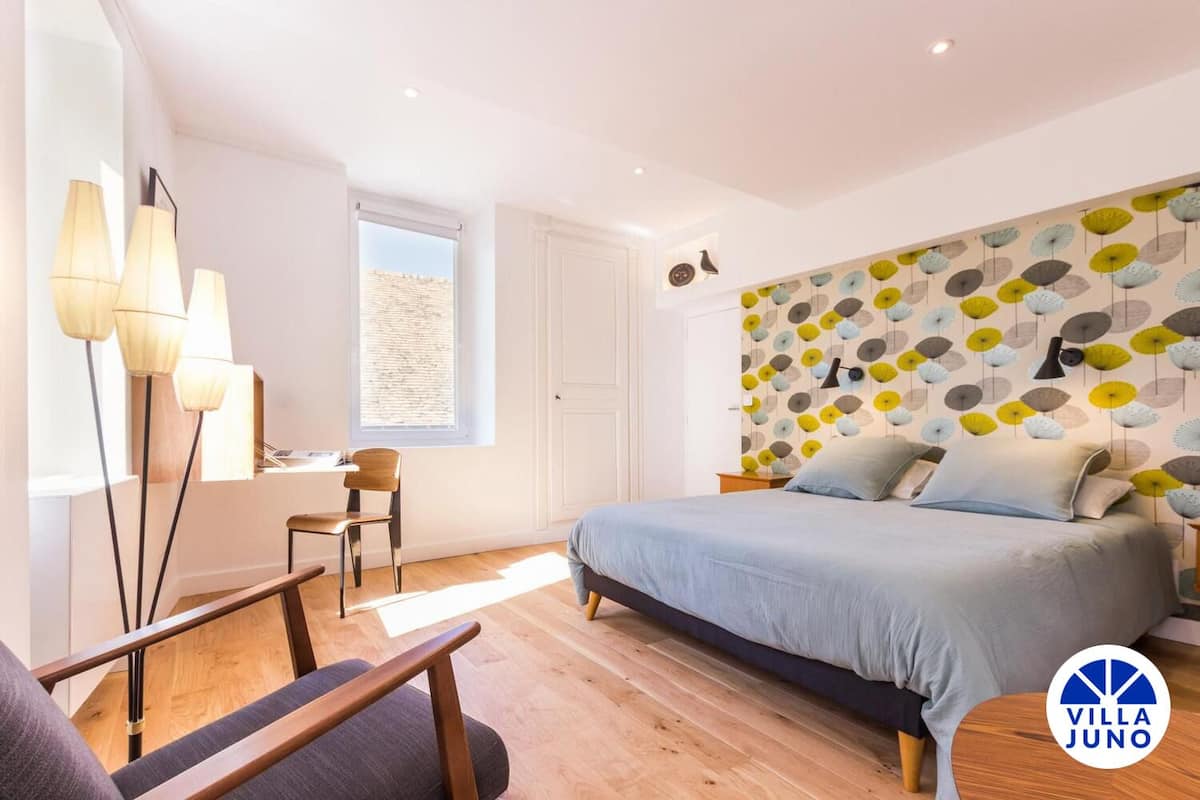 A modern bedroom with a bed, a desk, and a chair. There is a window with blinds, a wall with a patterned wallpaper, and a ceiling light.