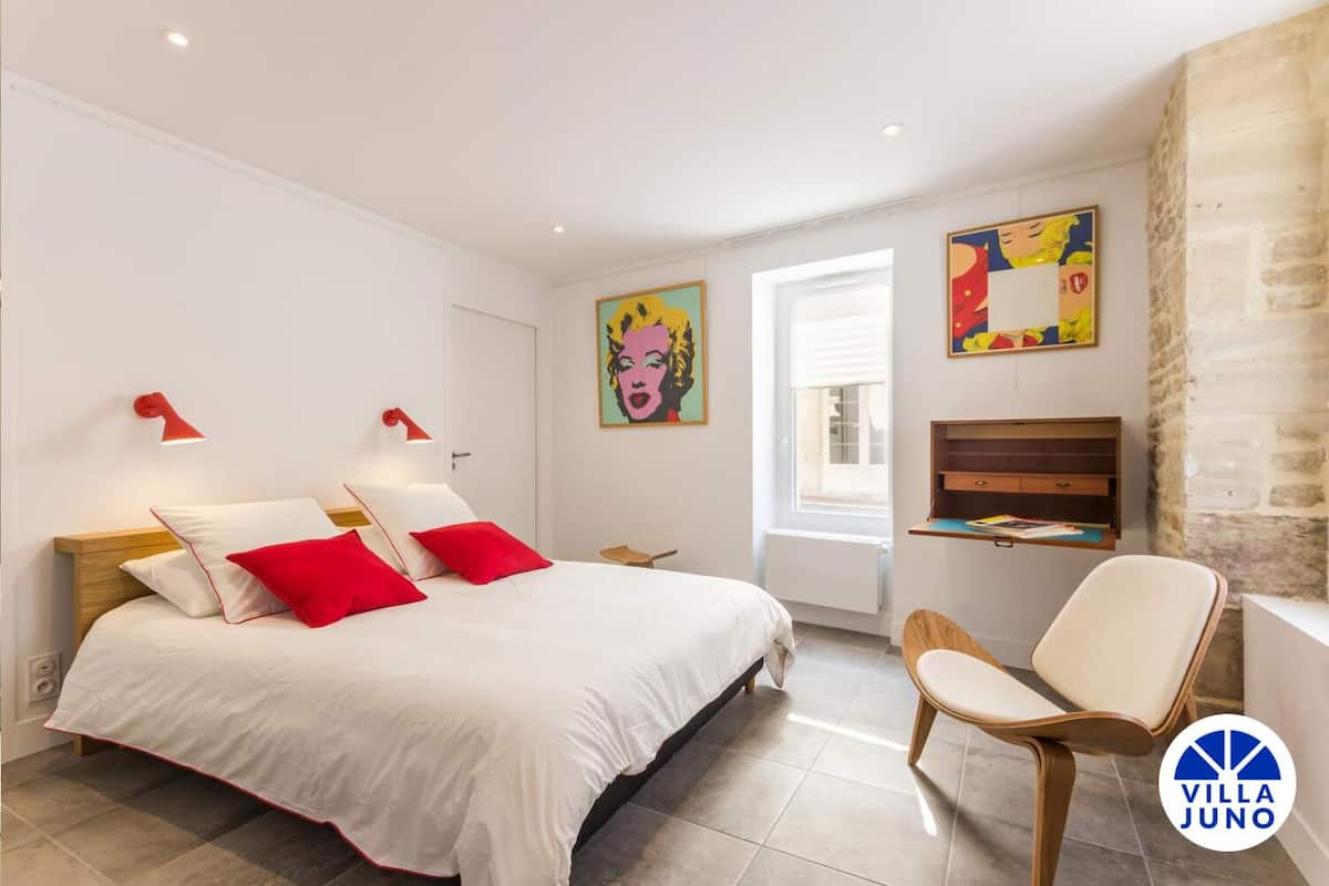 A bedroom with a bed, two red pillows, a chair, a desk, and two wall-mounted lamps.