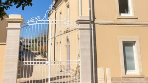 A gated entrance with a decorative metal gate, a stone pillar, and a window with shutters. - Villa Juno (Saint-Aubin-sur-Mer)