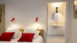 A bedroom with a bed, red and white pillows, a wooden headboard, a bedside table, a wall-mounted lamp, a mirror, and a bathroom with a sink and toilet. - Villa Juno (Saint-Aubin-sur-Mer)