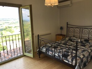 A bedroom with a metal bed frame, a quilted bedspread, a nightstand with a lamp, and a view through a sliding glass door to a balcony with a railing. - Charming 3 Bed Villa in Otricoli Stunnings Views (Otricoli)