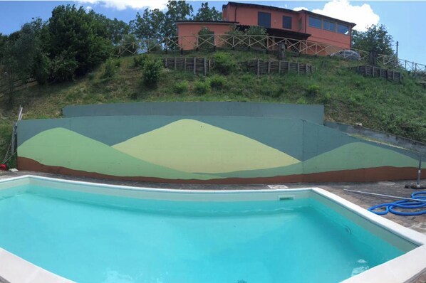 A swimming pool with a green and brown landscape mural, a red building in the background, and a grassy hill. - Charming 3 Bed Villa in Otricoli Stunnings Views (Otricoli)