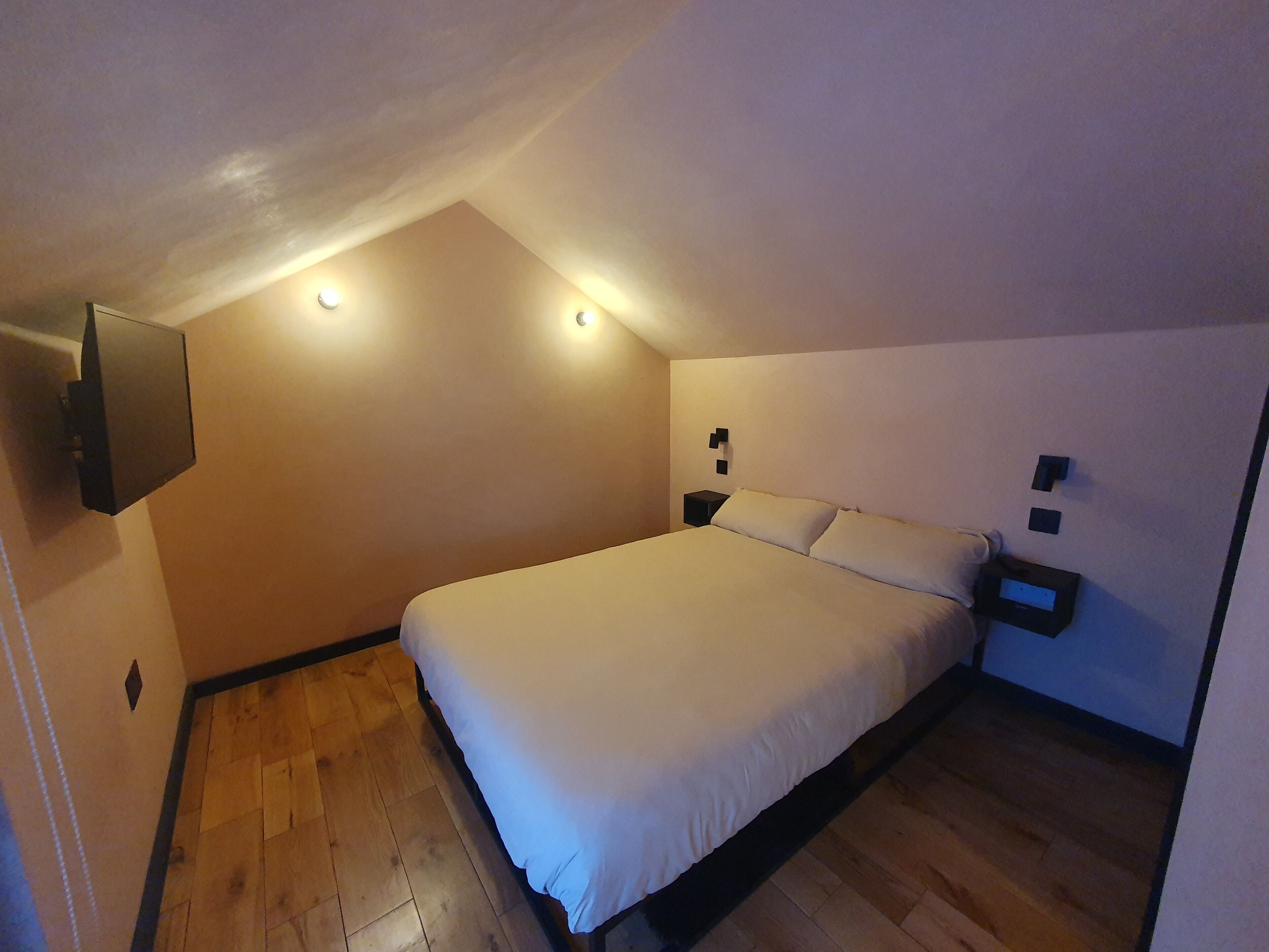 Ein Schlafzimmer mit einem großen Bett, Holzboden und einer schrägen Decke mit eingelassener Beleuchtung.