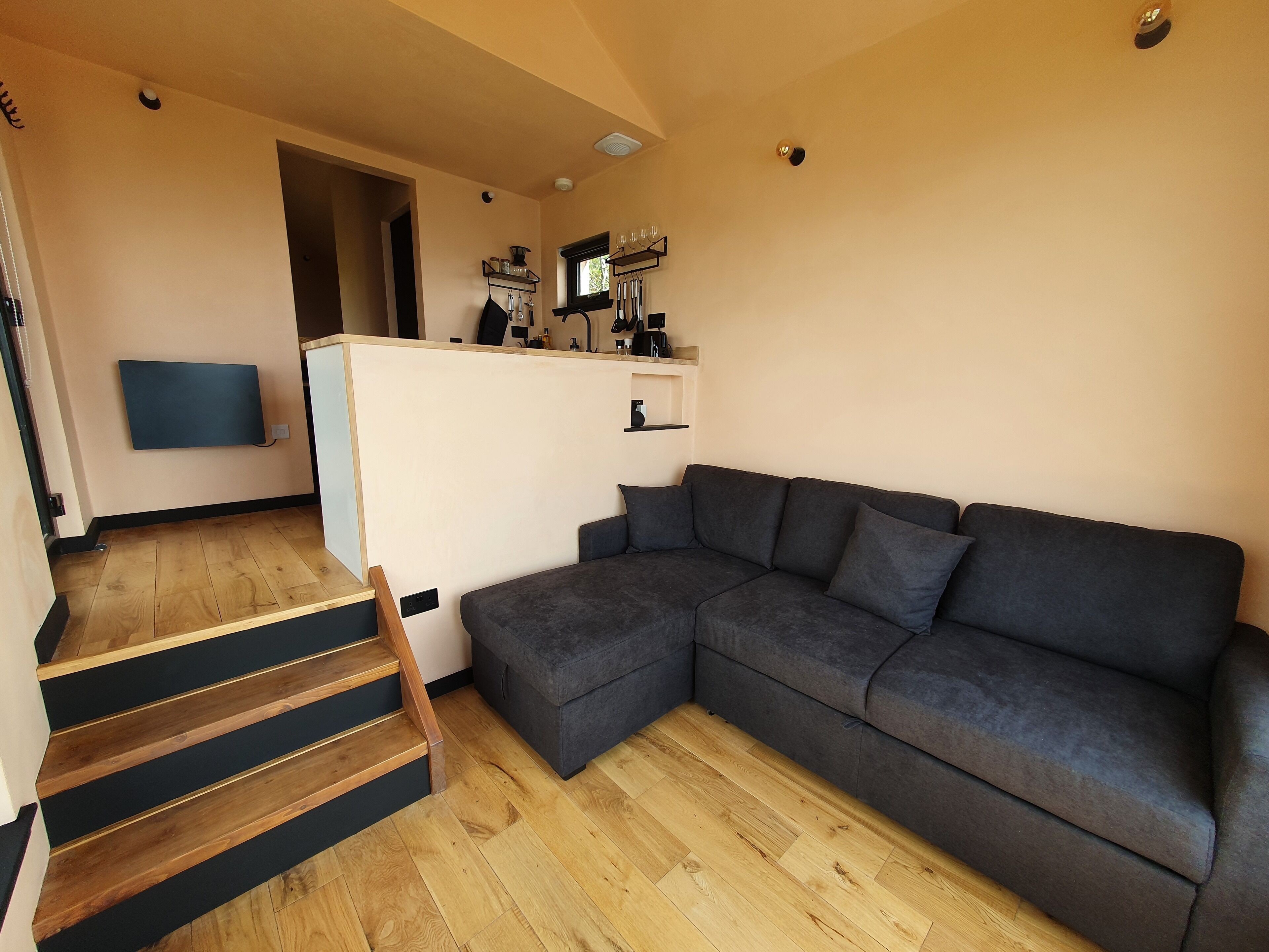 A modern living room with a grey sectional sofa, wooden flooring, and a kitchen area with a window.