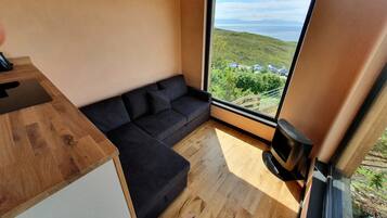 A compact living space with a sofa, a wooden table, and a large window offering a view of a hilly landscape.
