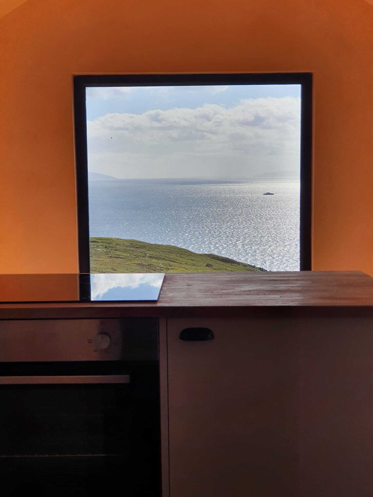Ett rum med utsikt över havet genom ett fönster, ett skrivbord i trä och ett litet bord med en bordsduk.
