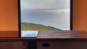 Ett rum med utsikt över havet genom ett fönster, ett skrivbord i trä och ett litet bord med en bordsduk.