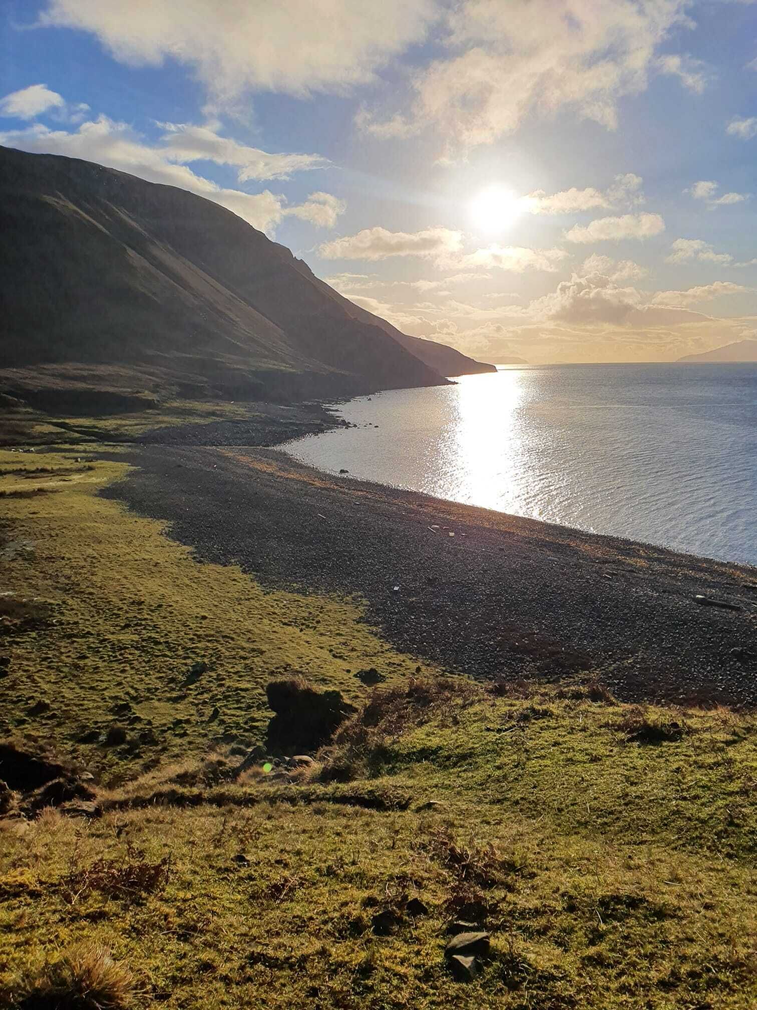 A coastal landscape with a pebbly beach, grassy areas, and a mountain in the background.