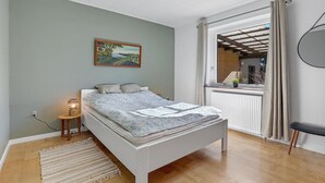 A bedroom with a bed, a bedside lamp, a painting on the wall, and a view of the outside through a window.