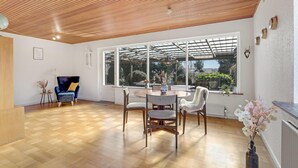A spacious room with a wooden ceiling, a large window with a view of the outdoors, a dining table with chairs, and a blue armchair.
