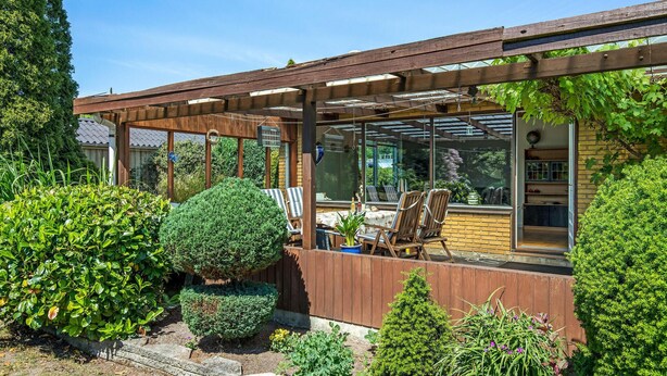 A covered patio area with a wooden pergola, a table with chairs, and a view of the garden through large windows.