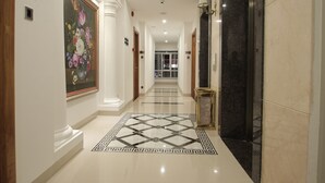A hotel hallway with a marble floor, a large floral painting on the left, and a black marble wall on the right. - Acnos Grand Hotel (Ho Chi Minh City)