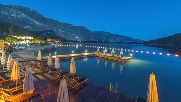 Ein Resort am Wasserufer mit Holzliegen, Sonnenschirmen und einer Anlegestelle, die in ruhiges Wasser führt.