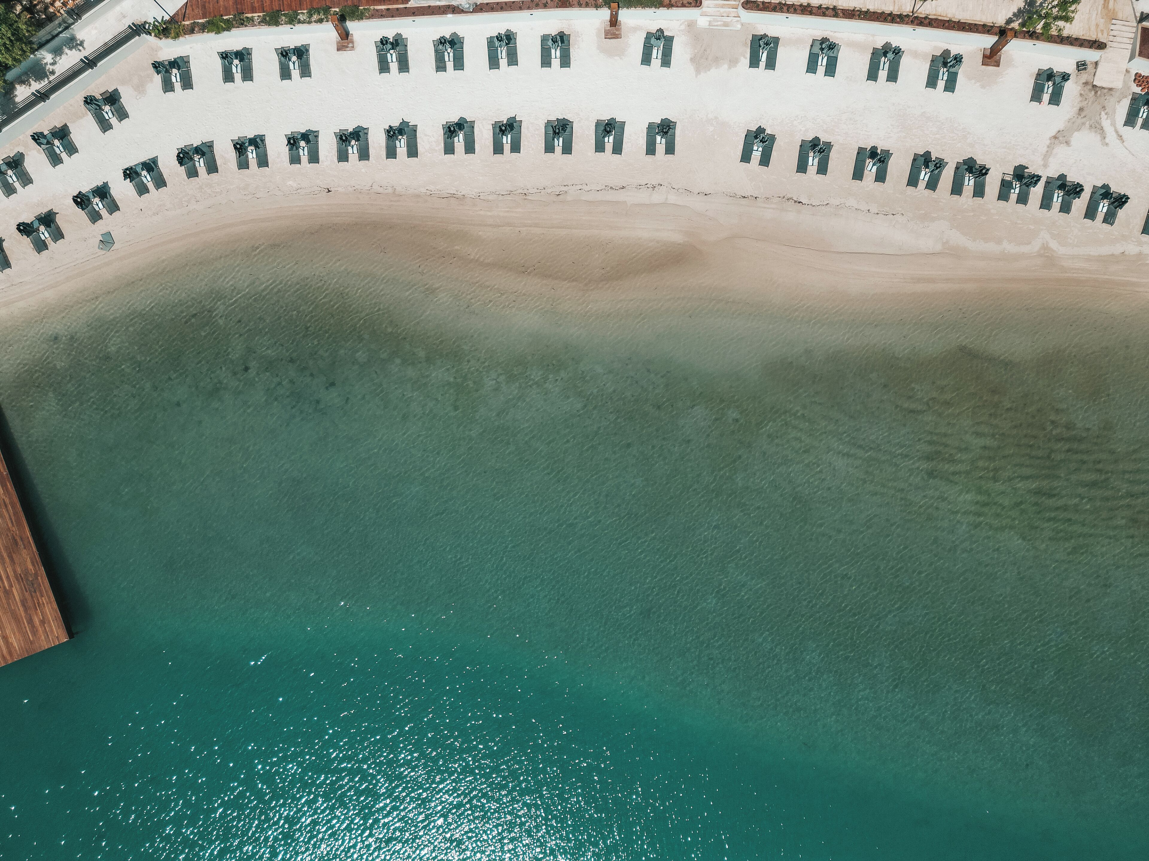 Vista aérea de una playa con hileras de sillas y sombrillas, una terraza de madera y un mar turquesa cristalino.