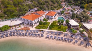 Luftaufnahme eines Küstenresorts mit mehreren Gebäuden, einem Strand mit Sonnenschirmen und einem Schwimmbad.