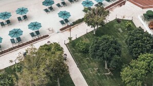 Eine Vogelperspektive zeigt einen Strand mit einer Holzstege, der sich in das türkisblaue Meer erstreckt, umgeben von Reihen von Strandsonnenschirmen und Liegestühlen.