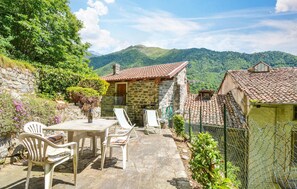 Une maison en pierre avec un toit de tuiles, une terrasse en pierre avec une table et des chaises, et une vue sur des collines verdoyantes en arrière-plan.
