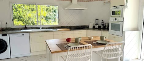 A kitchen with a large window, a washing machine, a dining table with chairs, and a microwave.