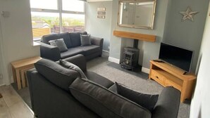 Una sala de estar con un sofá seccional gris, un mueble bajo para televisión de madera, un calentador pequeño y un ventanal con vistas al exterior.
