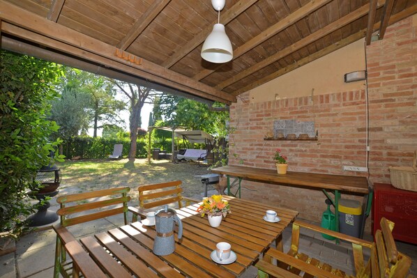 A wooden table with chairs, a bench, a hanging light fixture, and a brick wall with a window. - Casa Guna (Asciano)