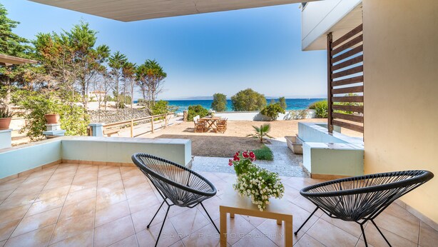 A patio with two black chairs, a small table, and a vase of flowers. It overlooks a beach and the sea. - Fluo Seaside Apartment (Aegina)
