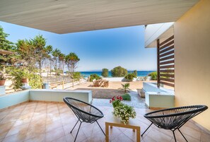 Un patio con dos sillas negras, una mesa pequeña y un jarrón de flores. Tiene vista a una playa y al mar.