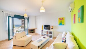 A modern living room with a sofa, coffee table, TV, and bookshelf. A fireplace is visible, and there is a view of the sea through large windows. - Fluo Seaside Apartment (Aegina)