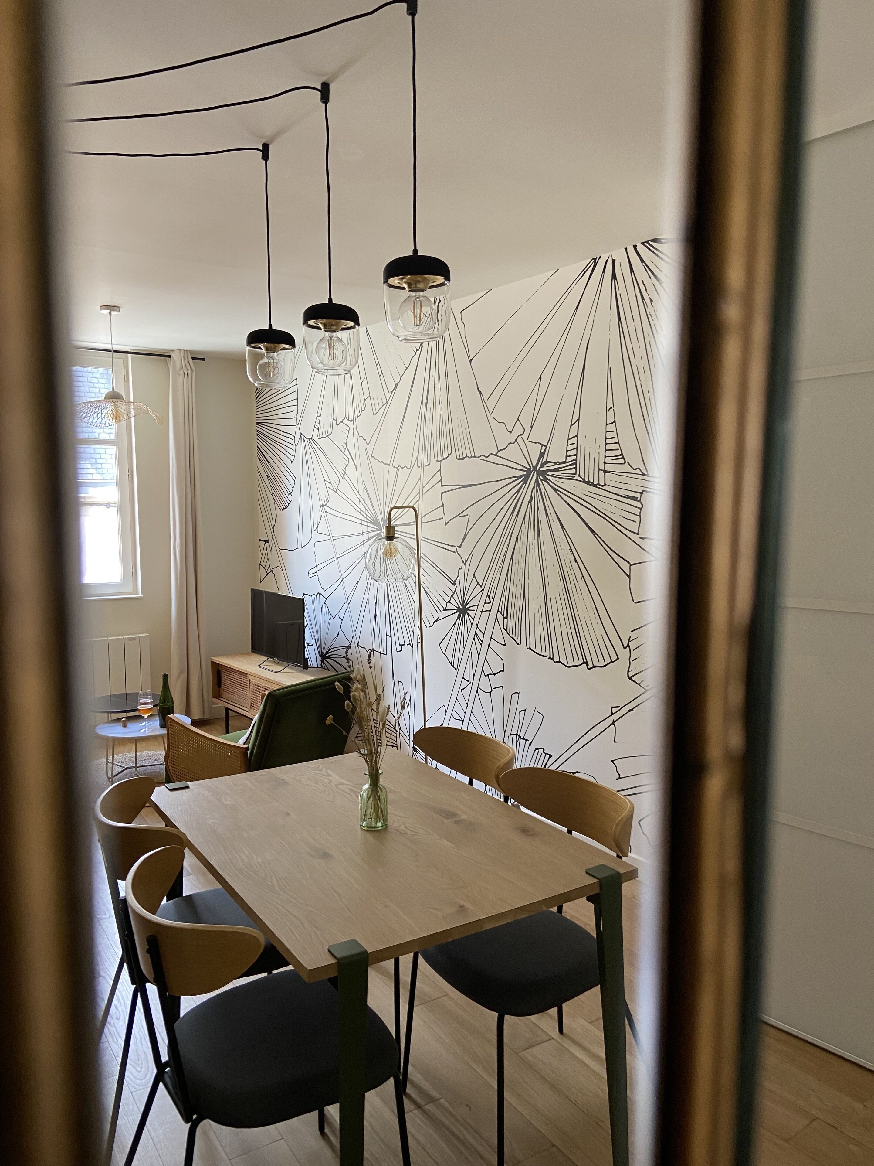 Ein Essbereich mit einem Holztisch, schwarzen und braunen Stühlen sowie Pendelleuchten. Im Hintergrund ist eine Wand mit geometrischem Muster zu sehen.