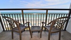 A balcony with two wicker chairs and a small table, overlooking a beach with people and umbrellas.