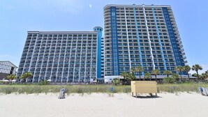 A large hotel with multiple floors and balconies, situated near a beach with palm trees.