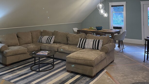 Een ruime woonkamer met een grote L-vormige zithoekbank, een salontafel en een gestreept vloerkleed. Een moderne kroonluchter hangt aan het plafond, en er is een eethoek met een tafel en stoelen zichtbaar op de achtergrond.