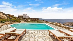 A pool area with lounge chairs and a stone-tiled floor, overlooking a mountainous landscape and the sea. - Villa Super Two (Mykonos)