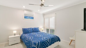 Une chambre à coucher comprenant un lit, une table de chevet, une lampe, un ventilateur de plafond, un appareil de climatisation, une fenêtre avec stores et un téléviseur posé sur un meuble.