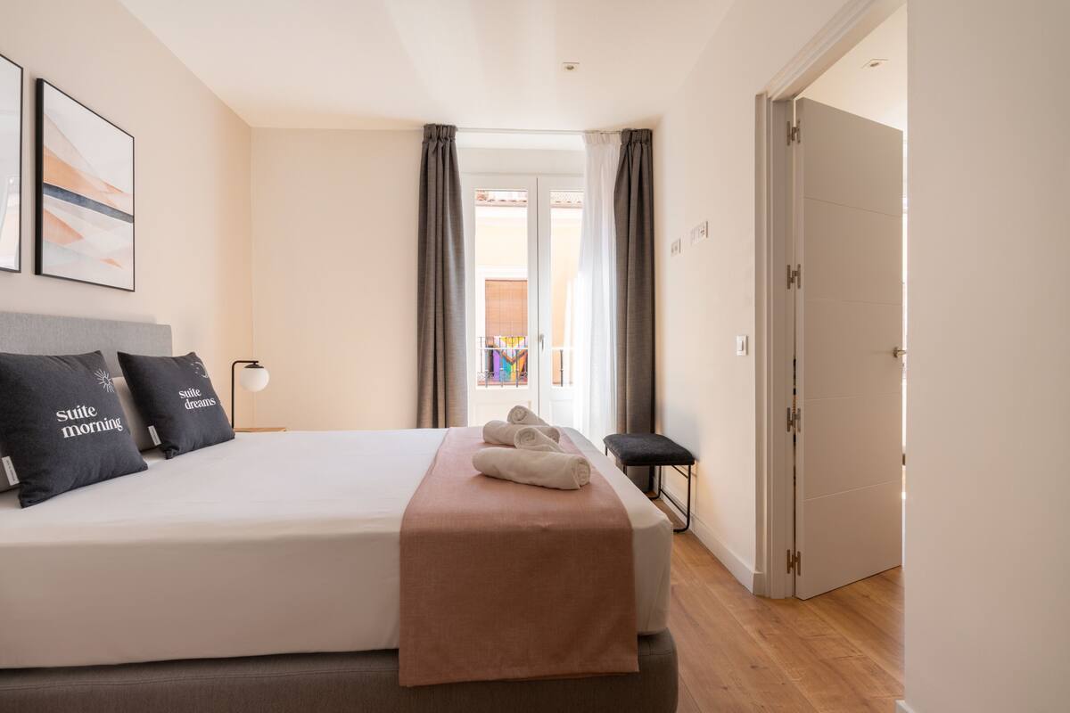 Una habitación de hotel con una cama grande, un banco, un asiento de banco, una mesita de noche y una puerta.