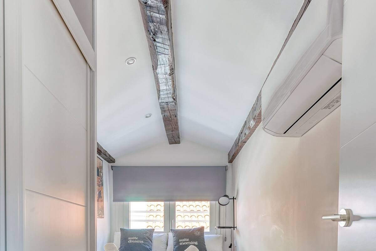 Un dormitorio con cama, suelo de madera, ventana con persianas y techo con vigas a la vista.