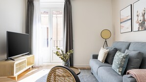 A modern living room with a grey sofa, a flat-screen TV on a wooden stand, a wicker chair, and a floor lamp. - Limehome Madrid C. de Fomento (Madrid)