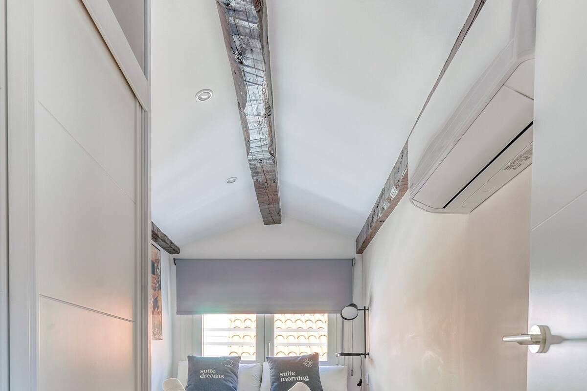 Un dormitorio con cama, almohadas, una mesita de noche, una ventana y suelo de madera.
