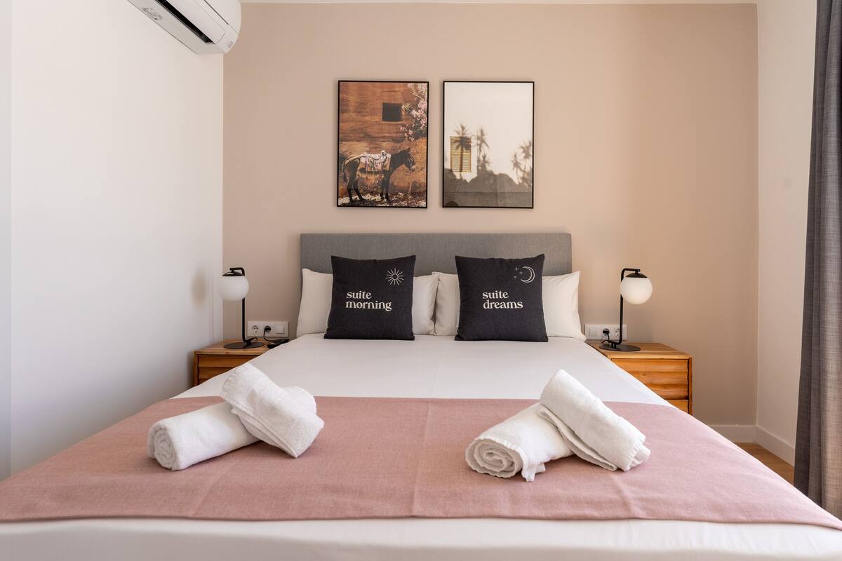 Una habitación de hotel con una cama, dos almohadas, dos mesitas de noche, un revistero y dos cuadros en la pared.