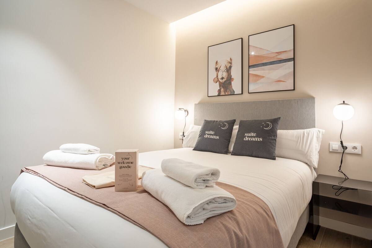 Una cama bien hecha con toallas dobladas, una mesita de noche con lámpara y dos cuadros en la pared.